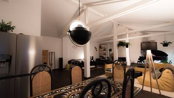 A modern living room with a dining area, featuring a large black bowl hanging light fixture, a piano, and a sofa.