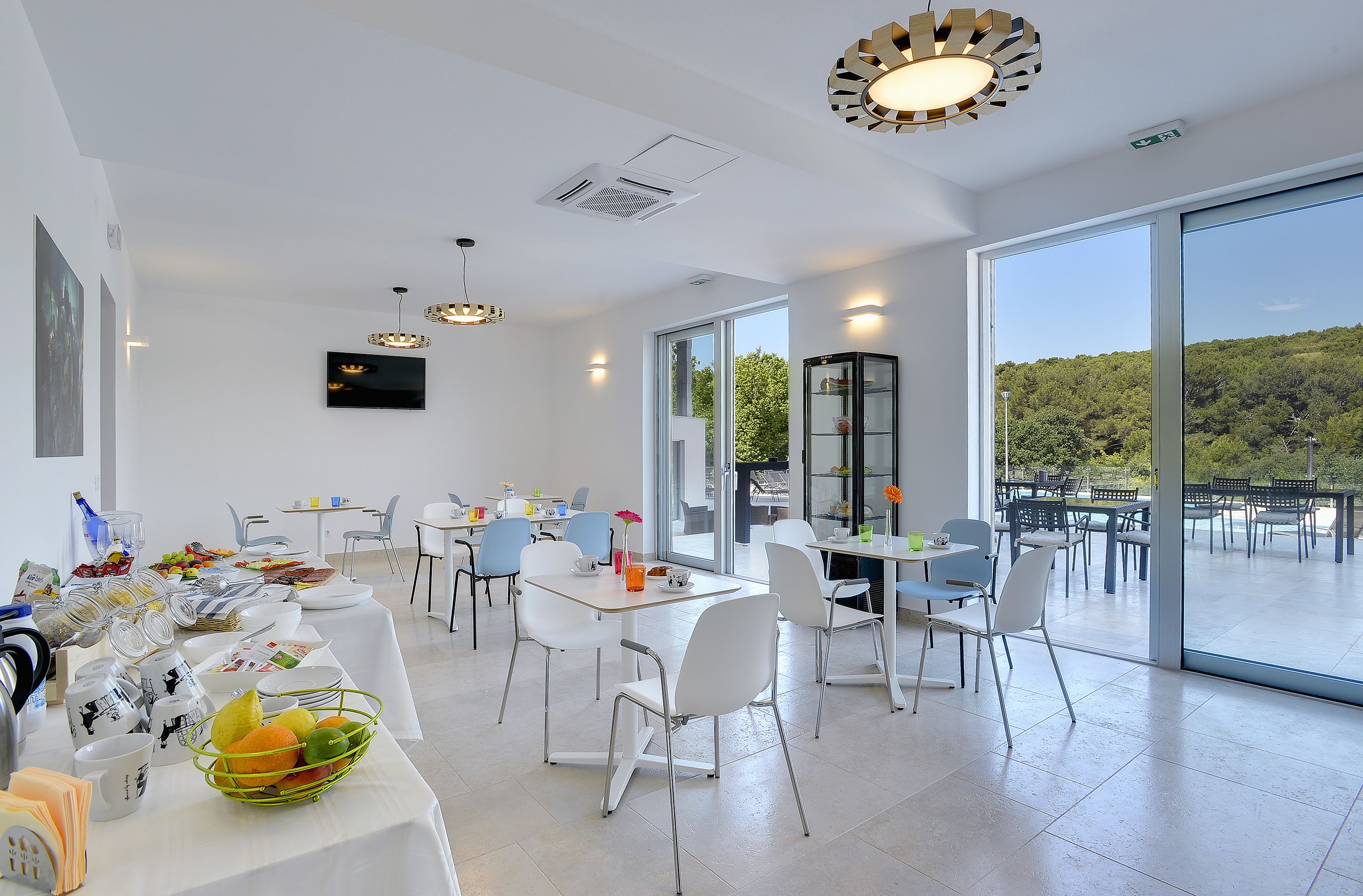 A modern dining area with white furniture, a large window offering a view of greenery, and a flat-screen TV mounted on the wall.