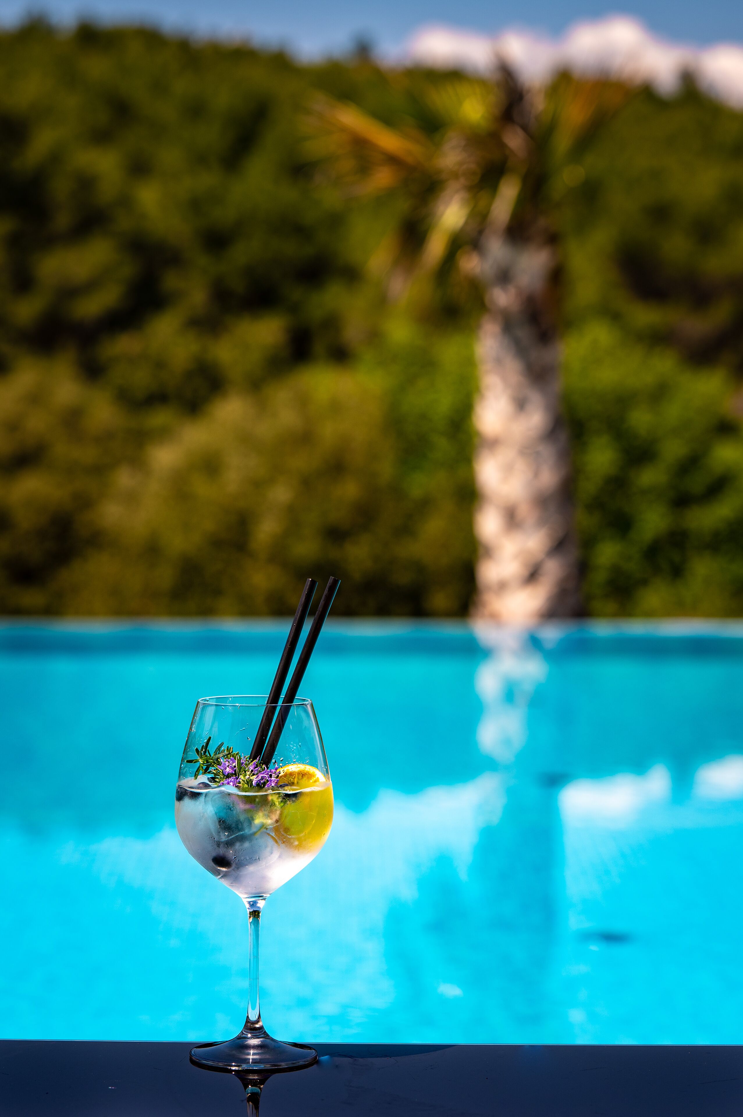 A clear glass with a lemon slice and a sprig of rosemary, placed on a reflective surface near a pool with a blurred tropical background.