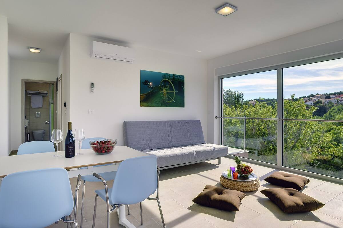 Uma sala de estar moderna com sofá, mesa de jantar, cadeiras e uma grande janela com vista para áreas verdes e casas.