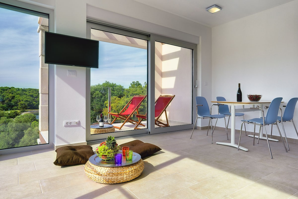 Quarto moderno com uma grande porta de correr de vidro que dá para uma varanda, televisão de tela plana na parede, mesa redonda com cesta trançada e cadeiras azuis ao redor de uma mesinha.