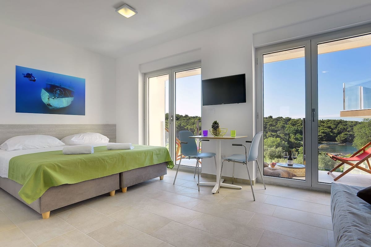 Quarto com cama, escrivaninha, cadeira, televisão e varanda com cadeira.