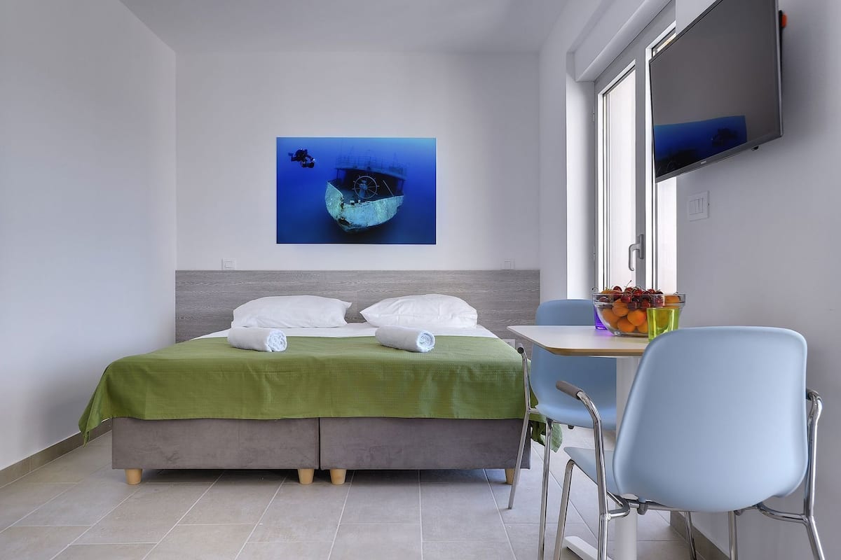 Quarto de hotel com cama, escrivaninha, cadeira, televisão e um quadro na parede.