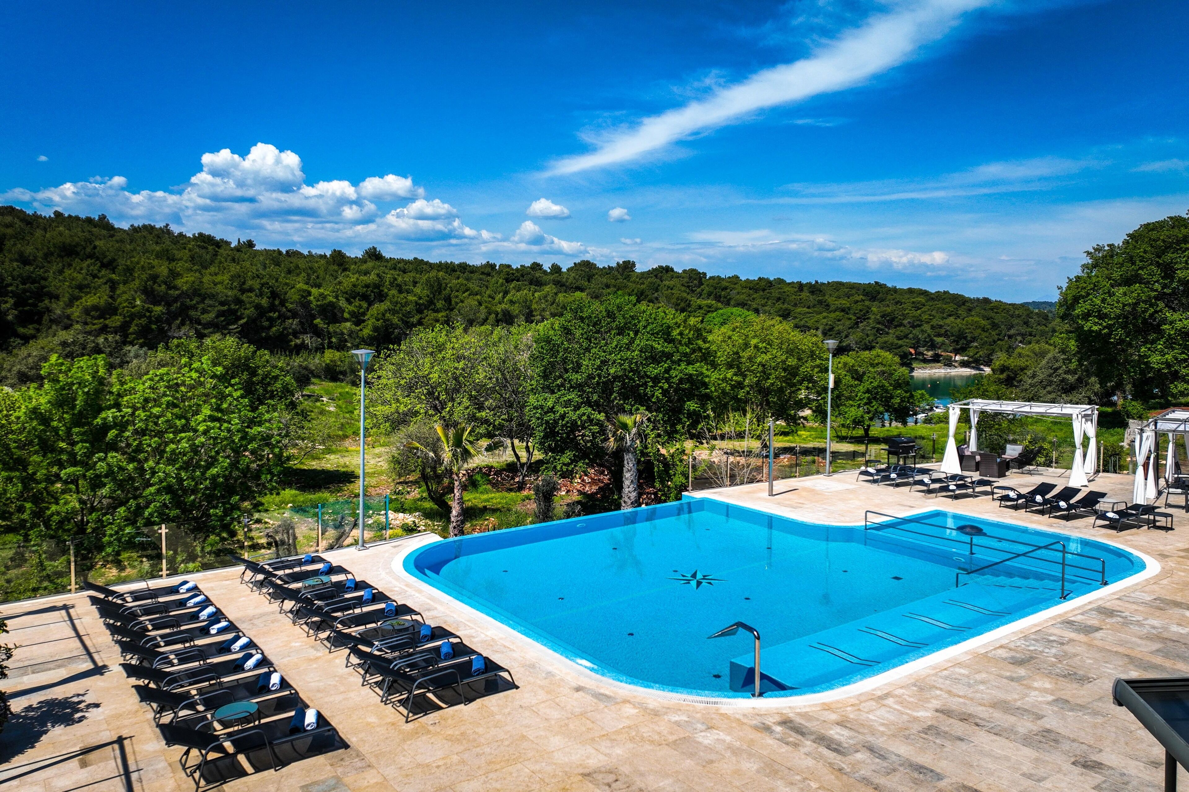 Ein Swimmingpool mit Liegestühlen und einer Laube, umgeben von Bäumen und blauem Himmel.