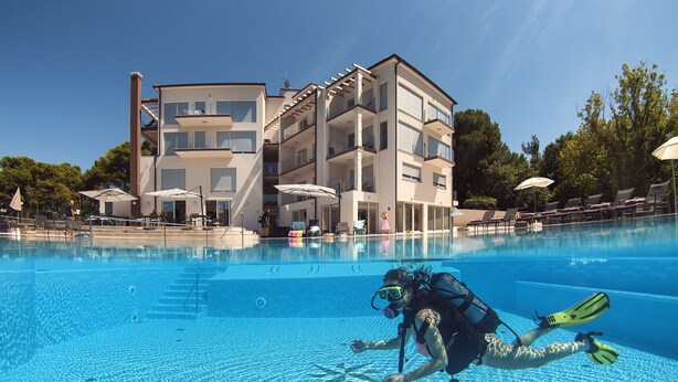A swimming pool with a diver in the water, surrounded by a modern building with balconies and outdoor seating.