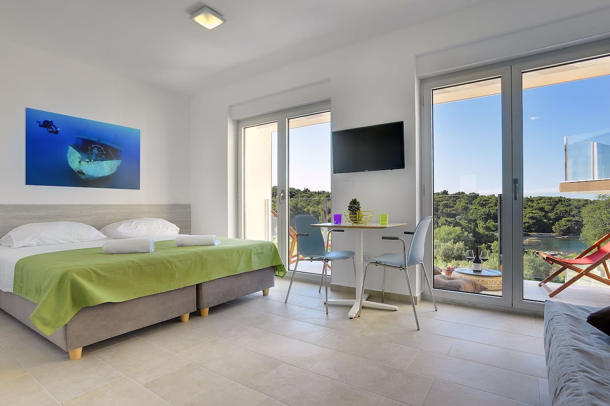 Quarto moderno com cama, escrivaninha e cadeira, TV fixada na parede, e uma janela grande com vista para vegetação e um corpo d'água.
