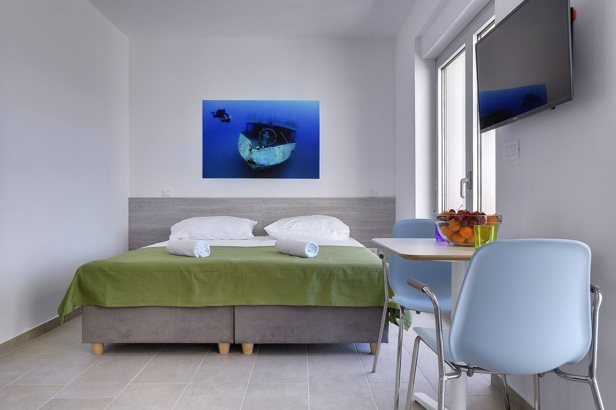 Quarto de hotel moderno com cama, escrivaninha, cadeira, televisão e um quadro de um naufrágio.