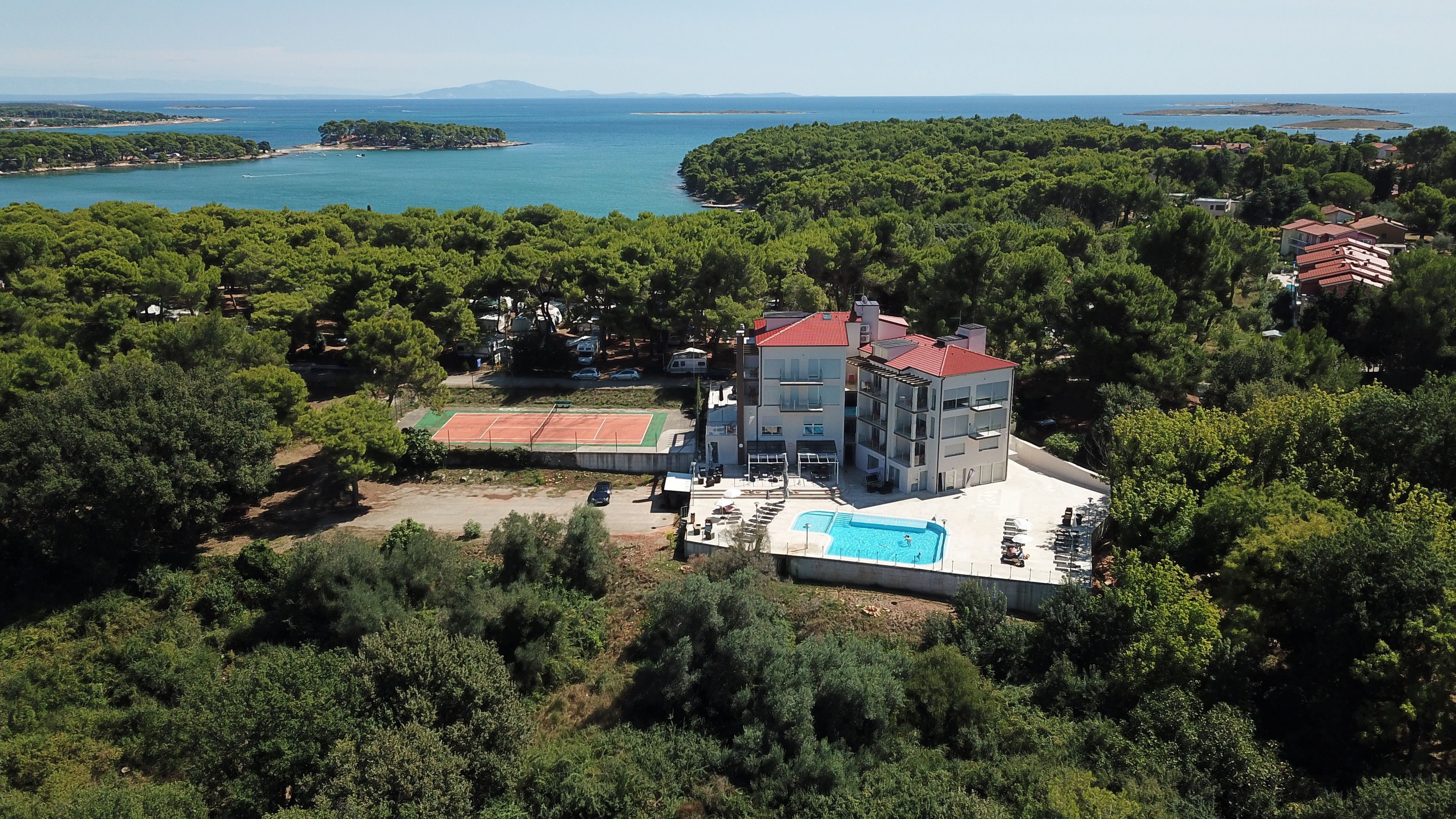 A multi-story building with a red roof, surrounded by dense greenery and a swimming pool.