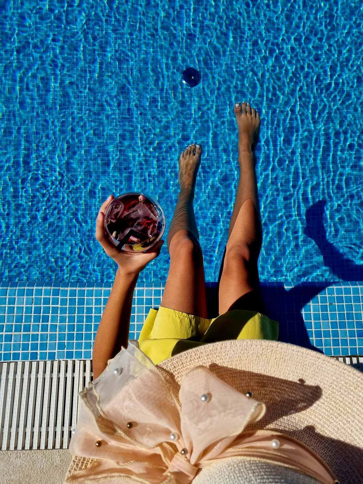 A person reclining by a pool with a drink in hand, wearing yellow shorts, and a white towel.