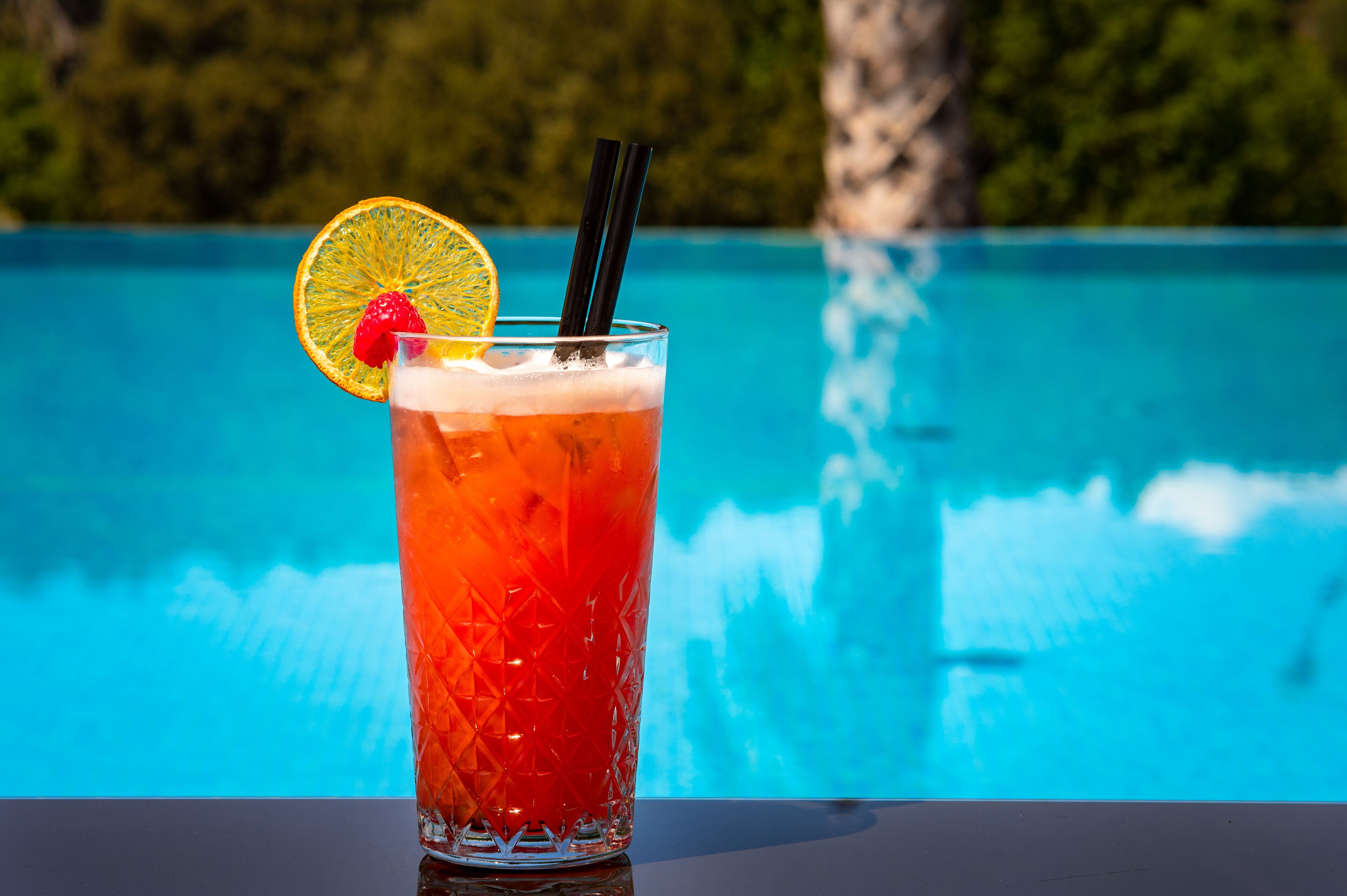A tall glass with a red beverage, garnished with a slice of orange and a strawberry, with black straws, placed in front of a pool.