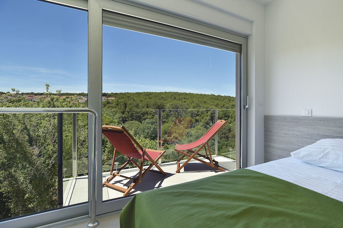 Uma varanda com uma cama, duas poltronas vermelhas e vista para uma área arborizada.