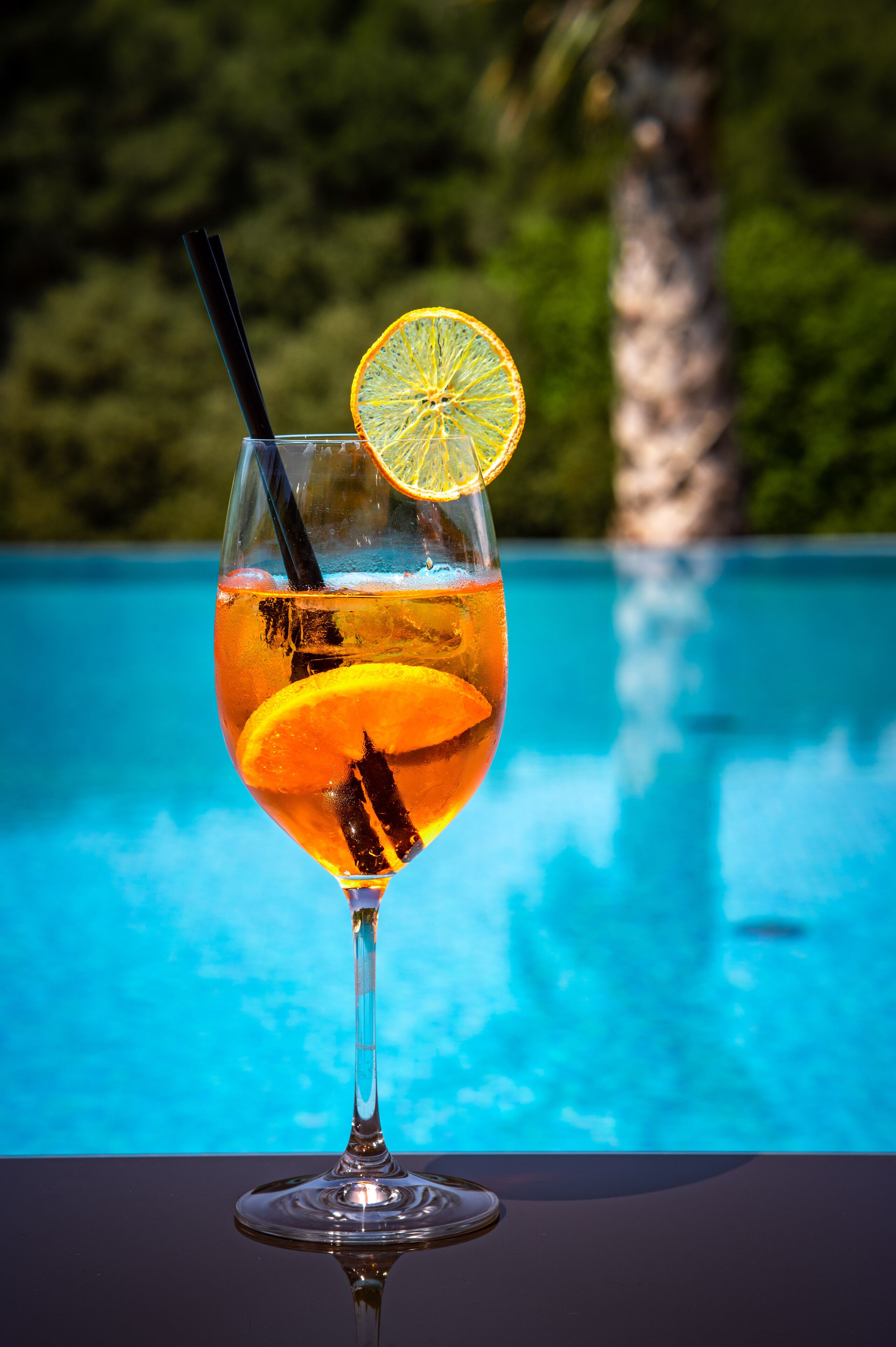 Un vaso de cóctel con una rodaja de naranja y una pajita, sobre una mesa junto a una piscina.
