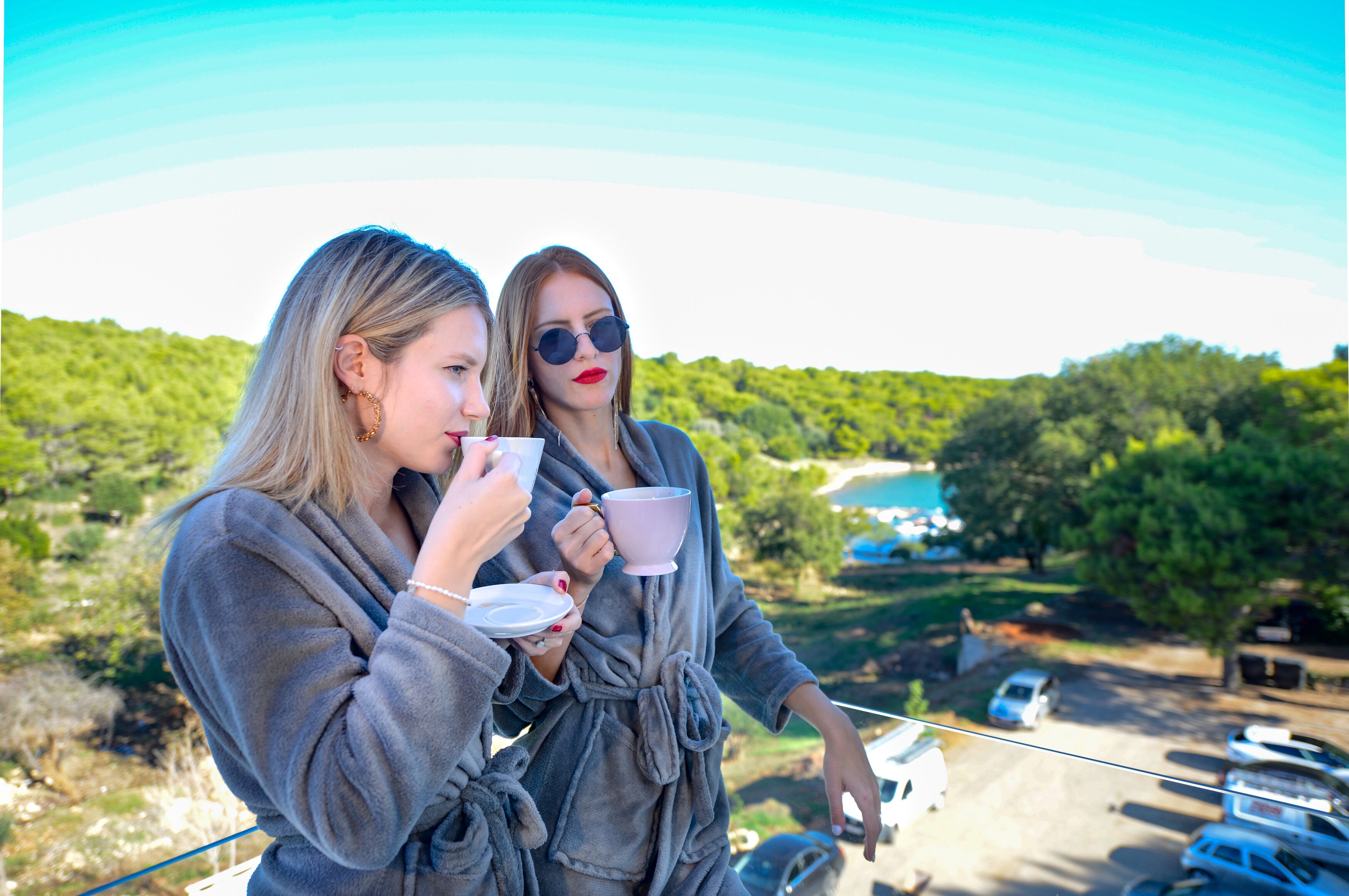Two people in robes on a balcony overlooking a parking area with trees and a body of water in the background.