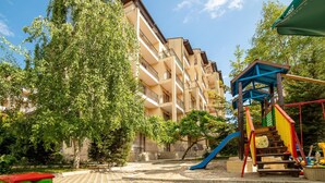Un parque infantil con tobogán y estructuras para escalar, rodeado de árboles y un edificio con balcones.