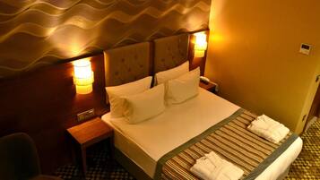 Quarto de hotel com cama, duas luminárias de cabeceira, mesa de apoio e uma cadeira.