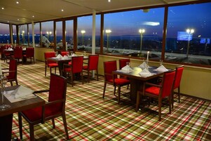 Un restaurante con sillas y mesas rojas, suelo a cuadros y amplias ventanas que ofrecen vista a la ciudad al atardecer.
