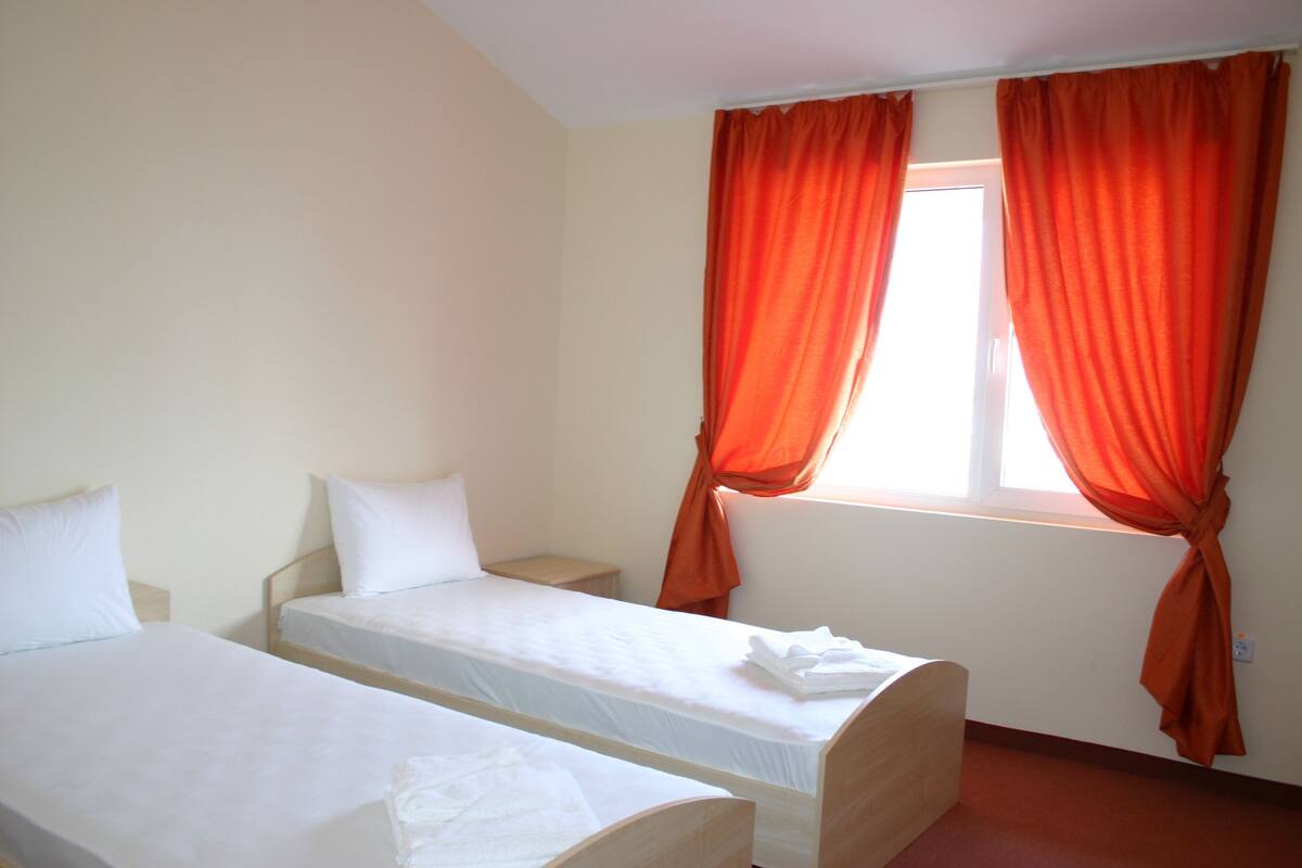 Ein Zimmer mit zwei Einzelbetten, einem Fenster mit orangenen Vorhängen und einem kleinen Nachttisch.