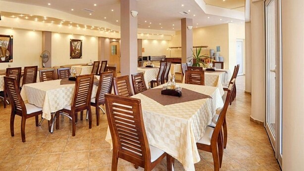 A dining area with multiple tables and chairs, a kitchen in the background, and a large window on the right. - Prestige City Apart Hotel (Sunny Beach)