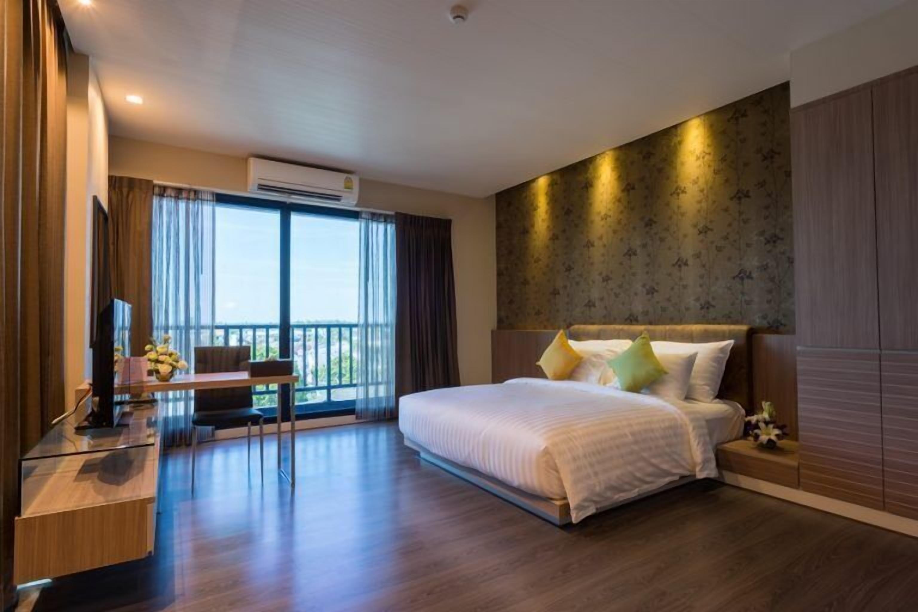Una habitación de hotel moderna con una cama grande, un escritorio con silla, un televisor y un balcón con cortinas.