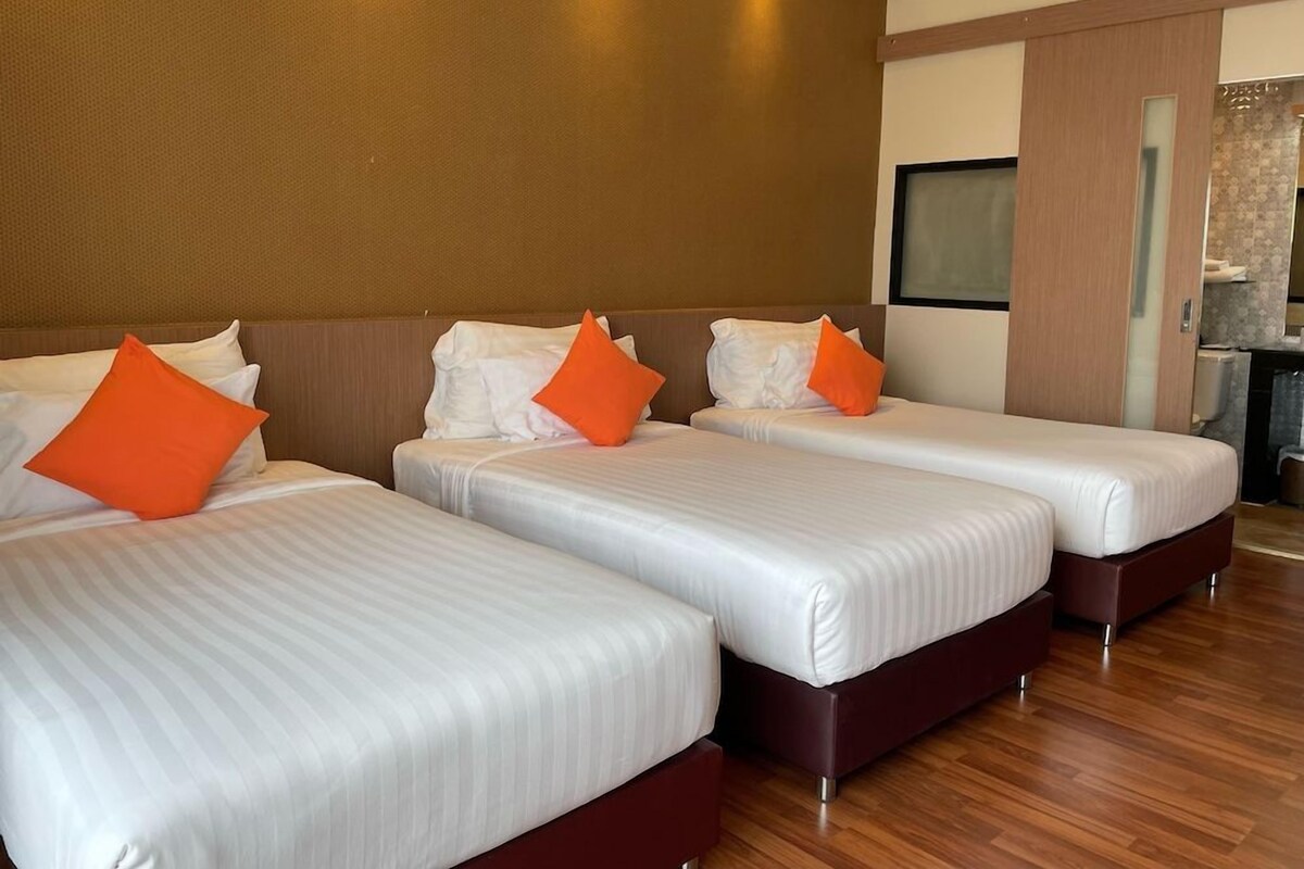 A hotel room with three beds, each with white bedding and orange pillows. The room has wooden flooring and a large mirror on the wall.