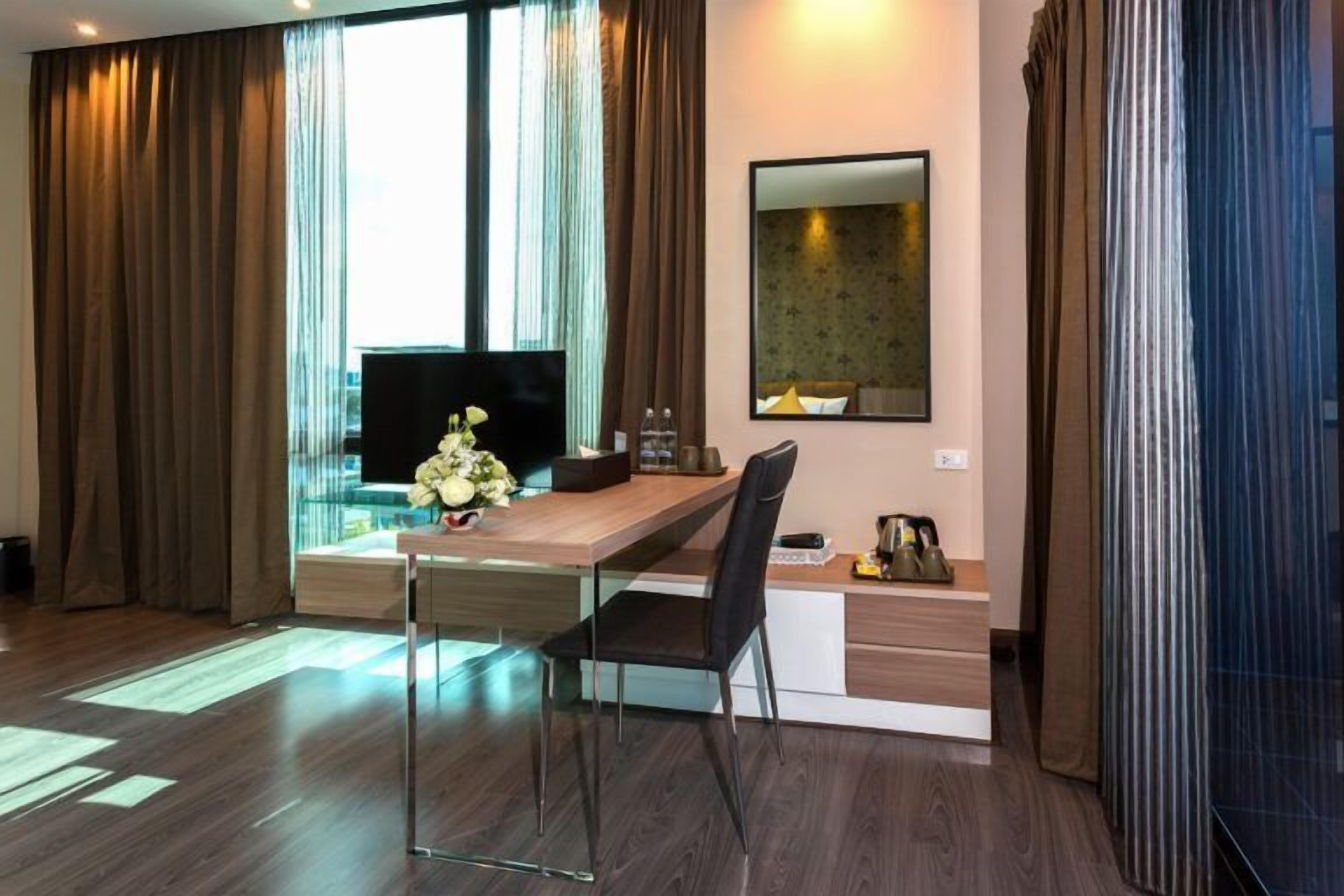 A modern hotel room with a wooden dining table, a black chair, a flat-screen TV, a vase with flowers, and a mirror on the wall.