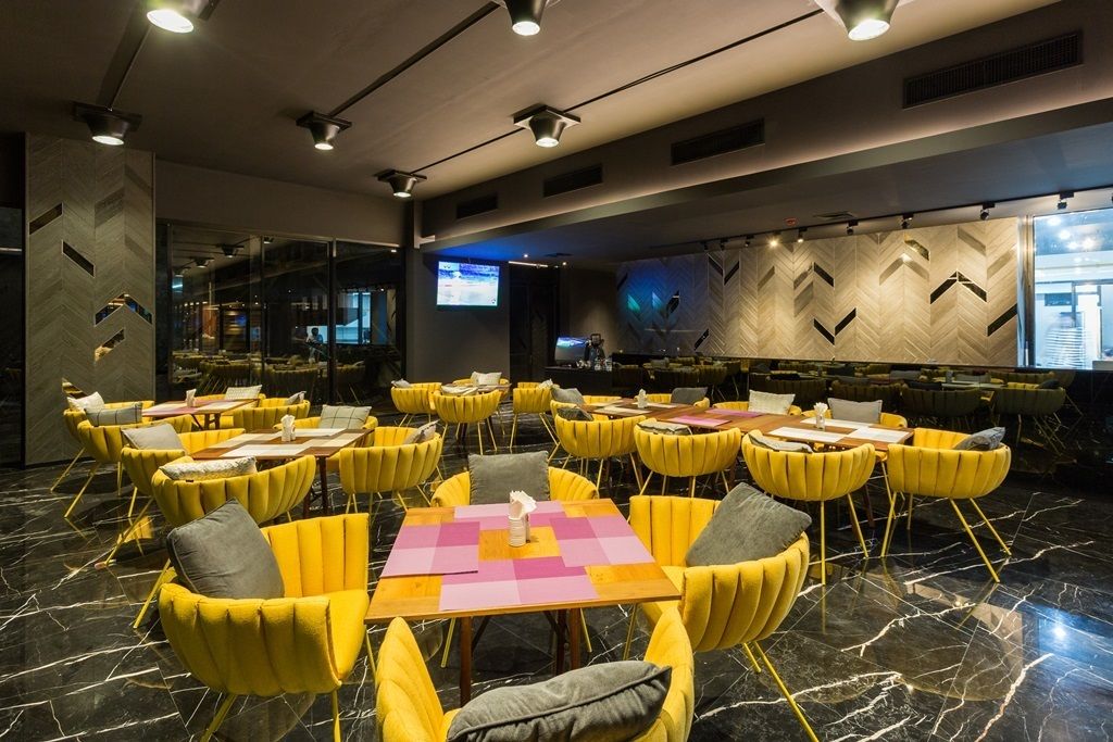 A modern dining area with yellow chairs and tables, a large screen, and a patterned wall.