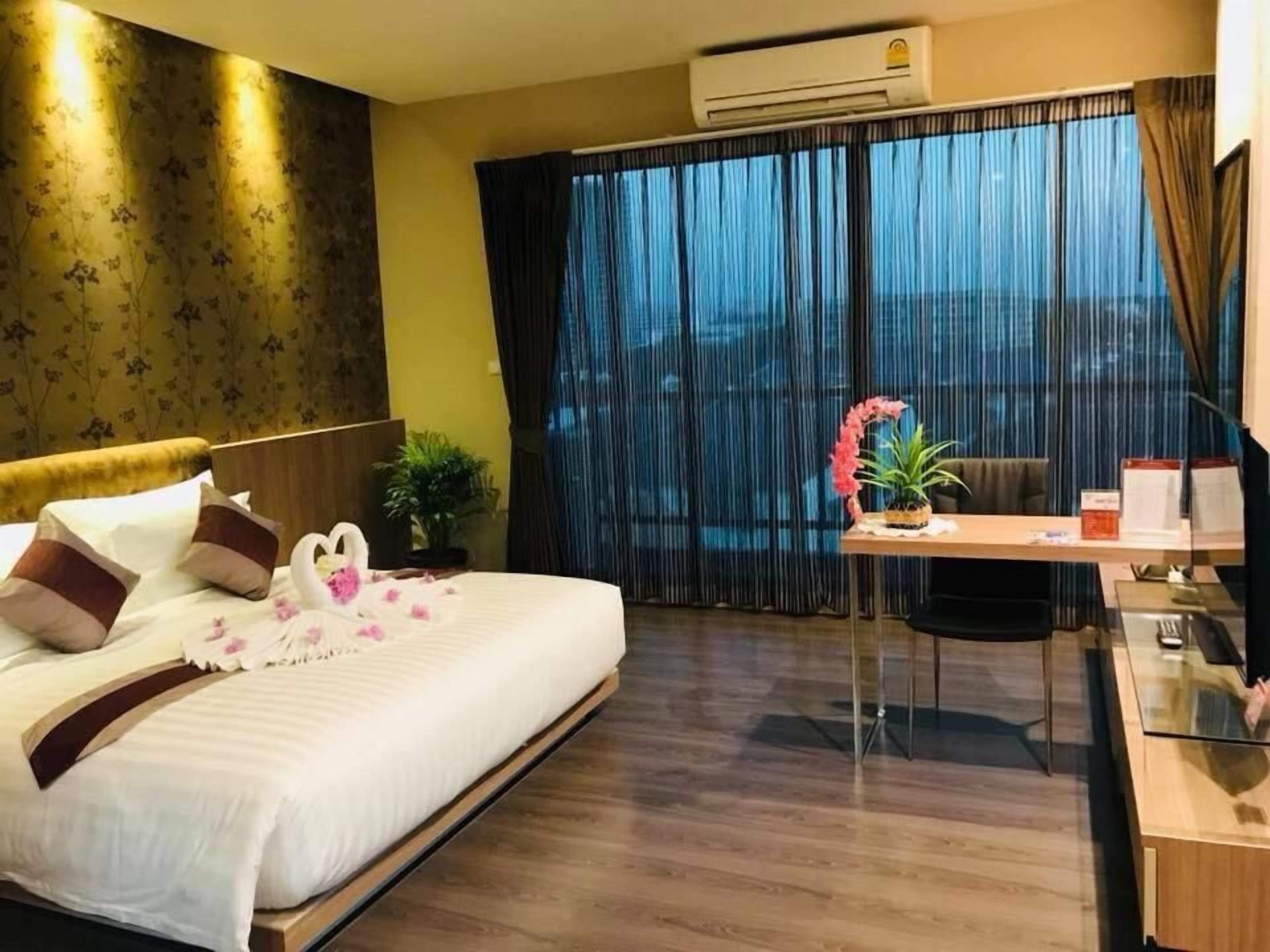 Een hotelkamer met een bed, een bureau, een stoel, een plant in pot en uitzicht buiten door grote ramen.