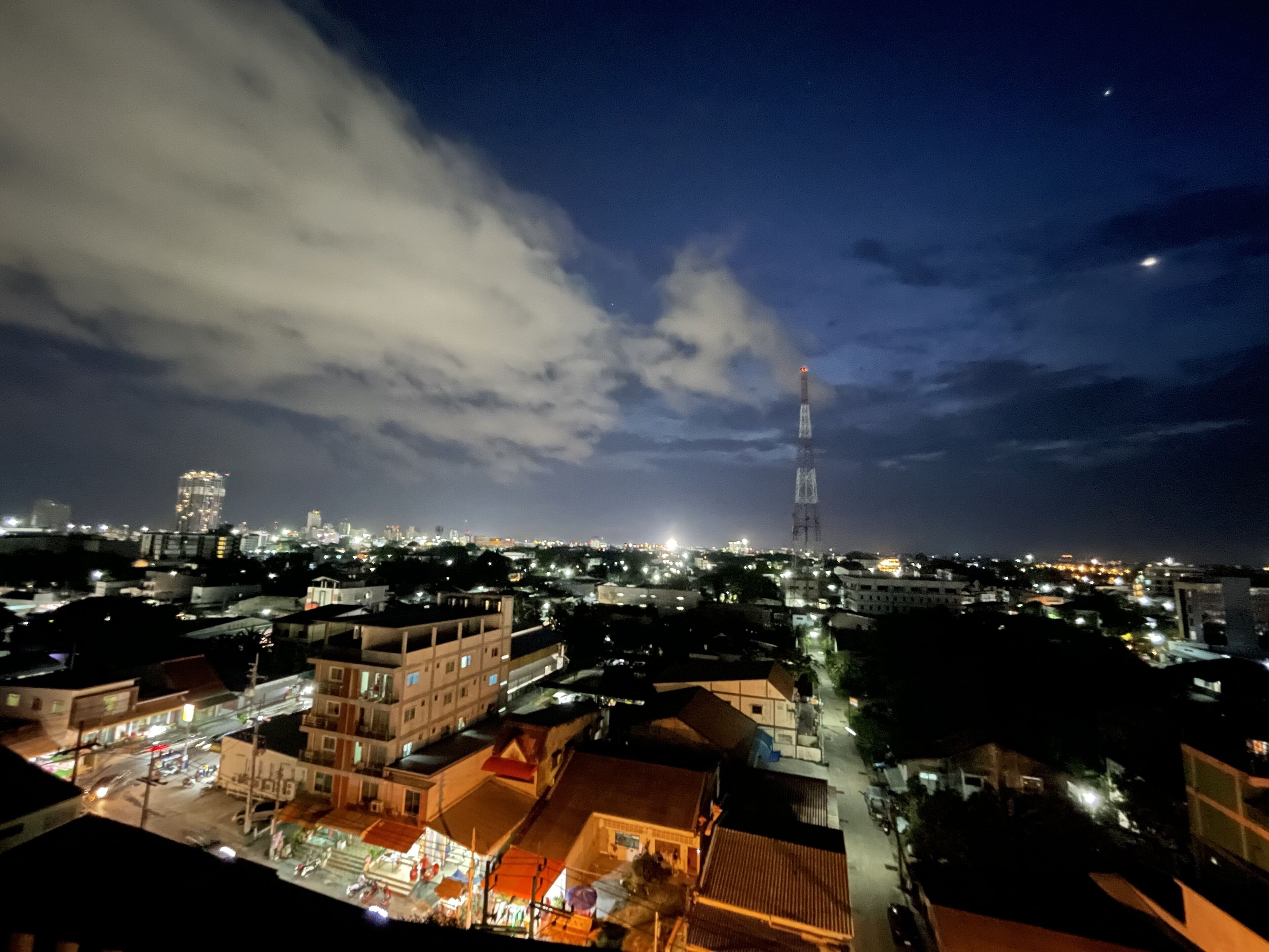 Un paisaje urbano nocturno con una torre destacada, varios edificios y una calle concurrida abajo.