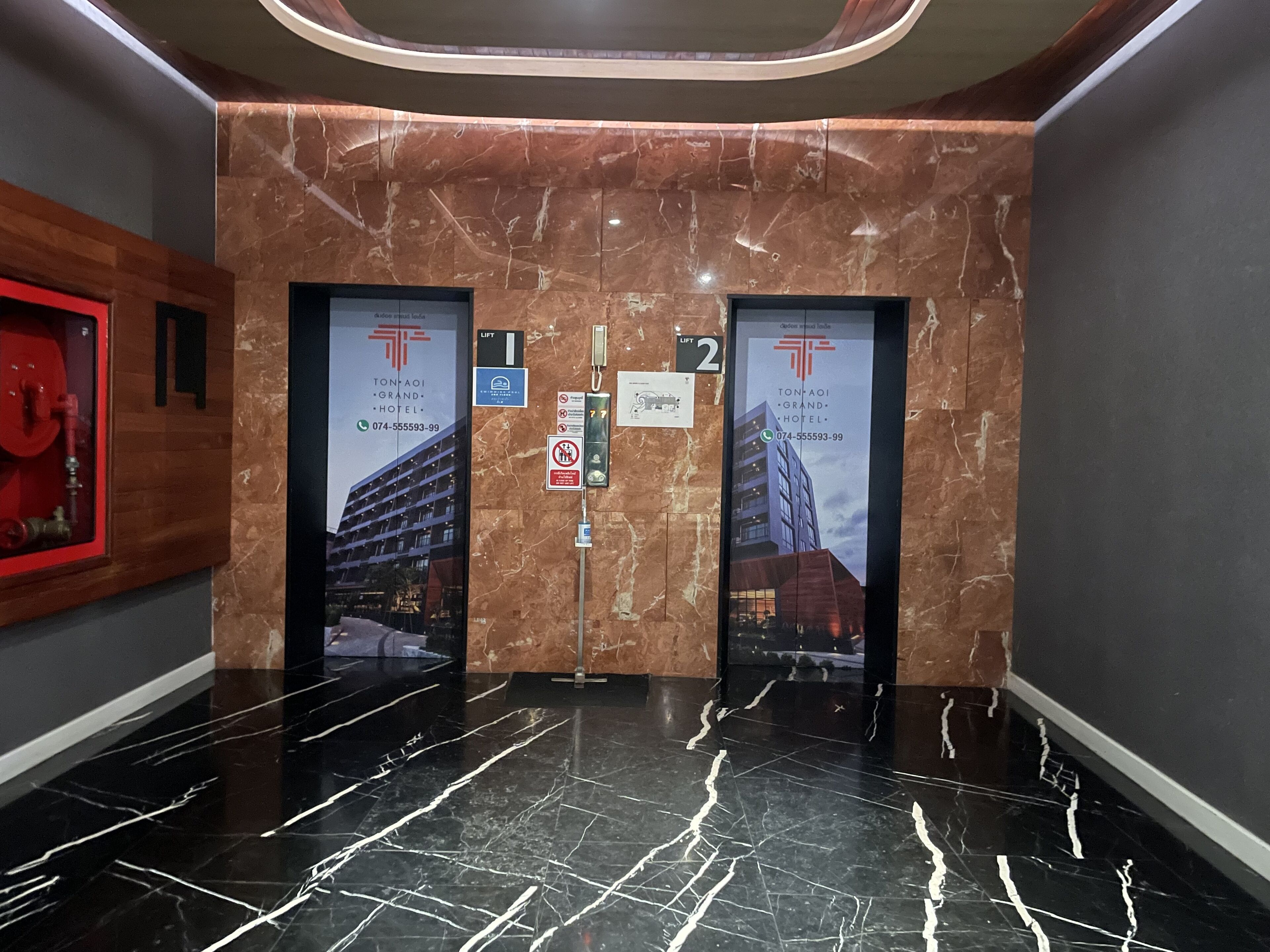 A modern elevator interior with marble walls and black marble floors. There are two elevators, each with a sign indicating 'Lift 1' and 'Lift 2'. The walls have a logo and contact information for 'Ton Paoi Grand Hotel'.