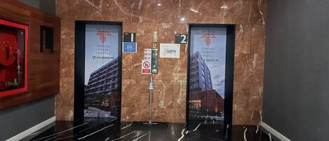 A modern elevator interior with marble walls and black marble floors. There are two elevators, each with a sign indicating 'Lift 1' and 'Lift 2'. The walls have a logo and contact information for 'Ton Paoi Grand Hotel'.