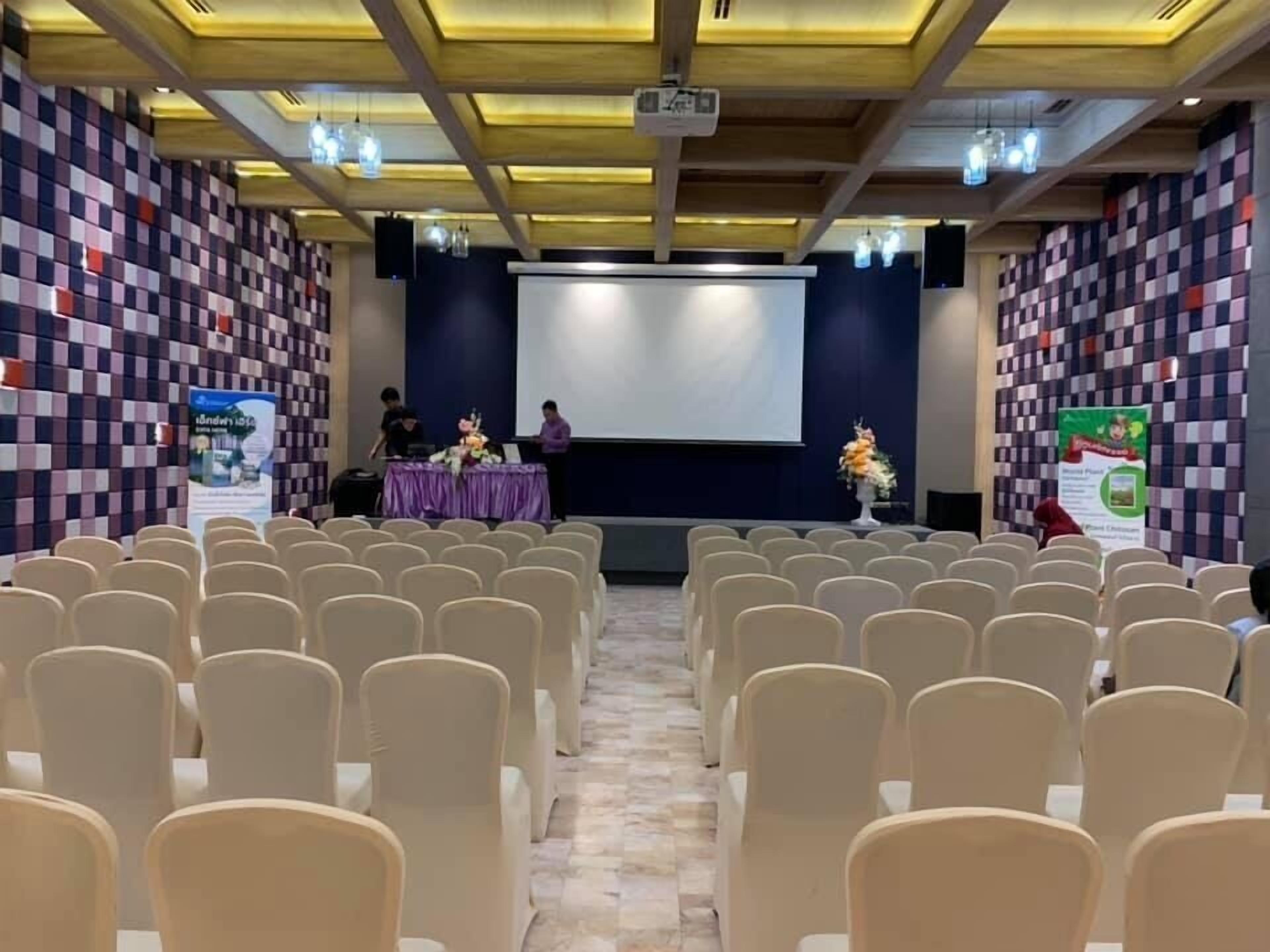 A conference room with rows of chairs, a stage with a projector, and two speakers.