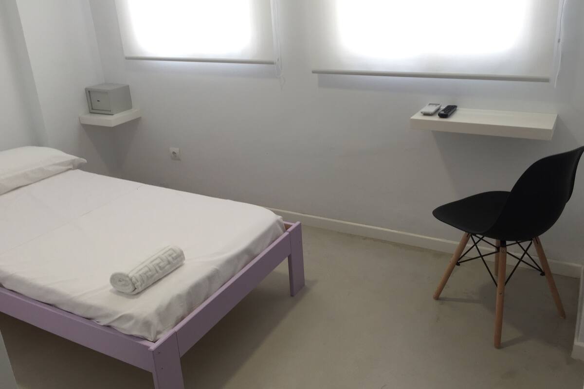 Un dormitori amb un llit individual, una cadira, una estant amb un telèfon i una taula petita.