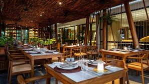 A restaurant with wooden tables and chairs, large windows, and indoor plants. - Anjali by Syphon (Siem Reap)