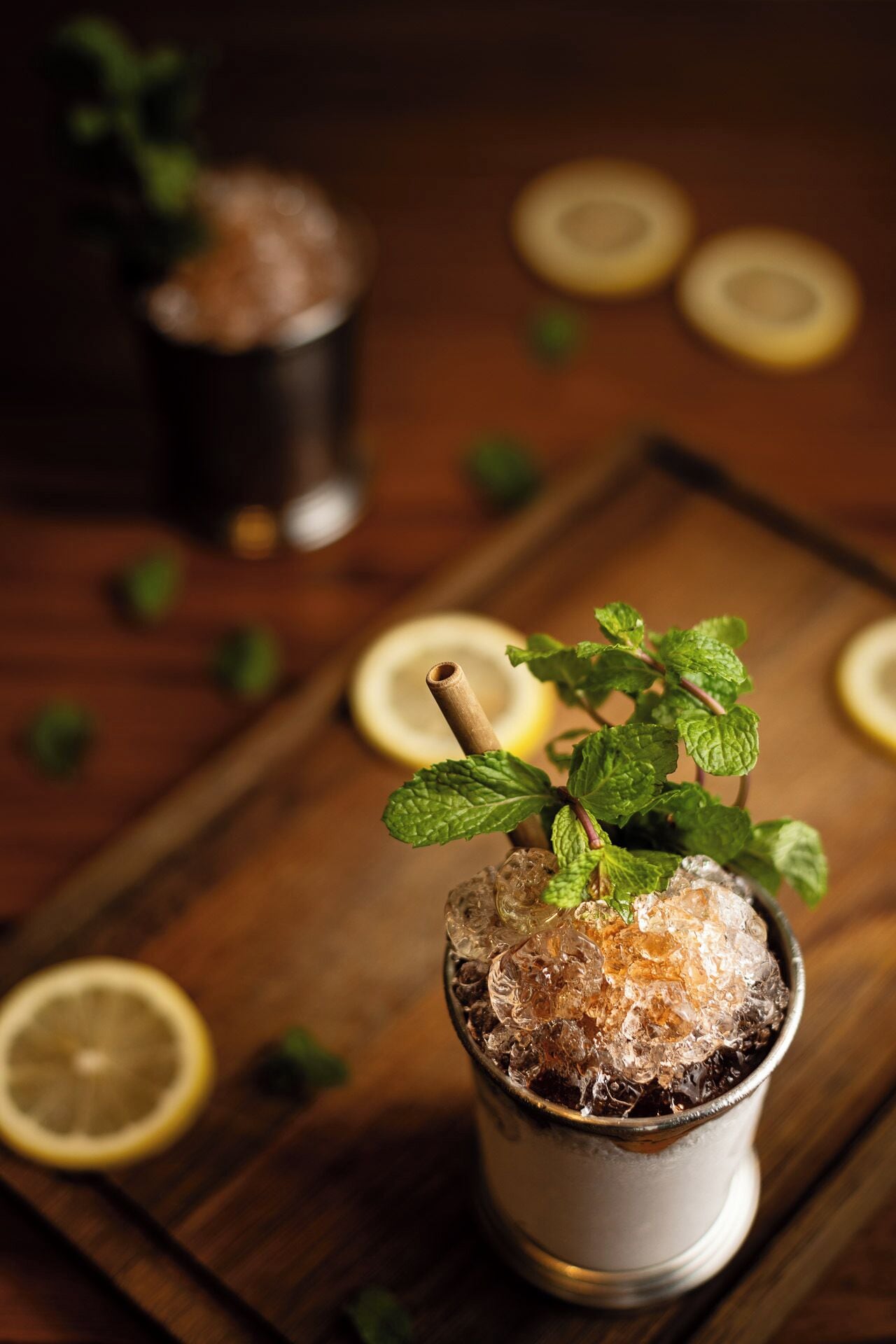 A glass of iced beverage with mint leaves and a straw, surrounded by lemon slices on a wooden surface.