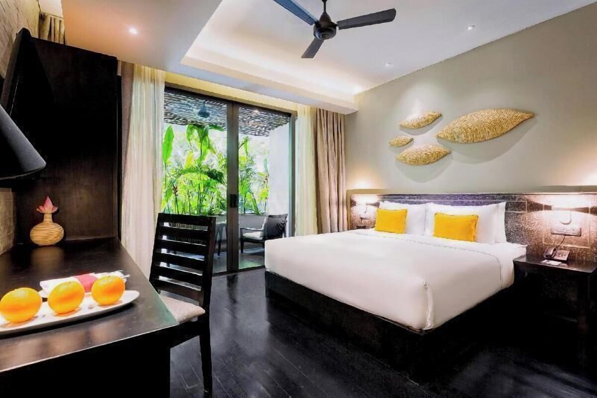 A hotel room with a bed, a desk with a lamp, a chair, and a view of greenery through the window.