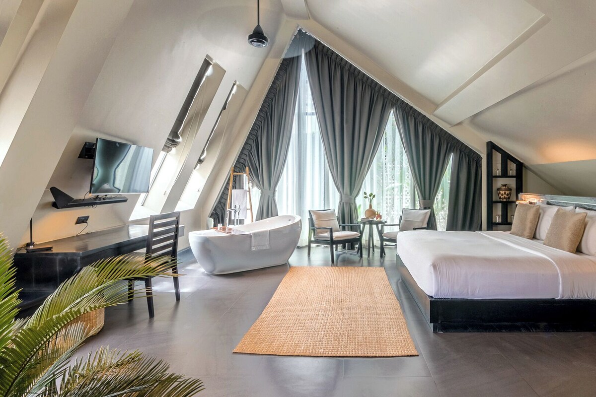 A modern hotel room with a large bed, a sitting area with a bathtub, and a TV.