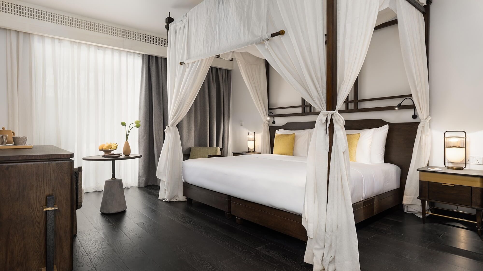 A hotel room with a four-poster bed, a bedside table with a lamp, a small table with a vase and fruit, and a wardrobe.