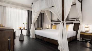 A hotel room with a four-poster bed, a bedside table with a lamp, a small table with a vase and fruit, and a wardrobe.