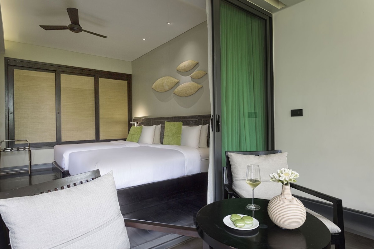 Una habitación de hotel con una cama, una silla, una mesa con un florero y un vaso, y una cortina verde.