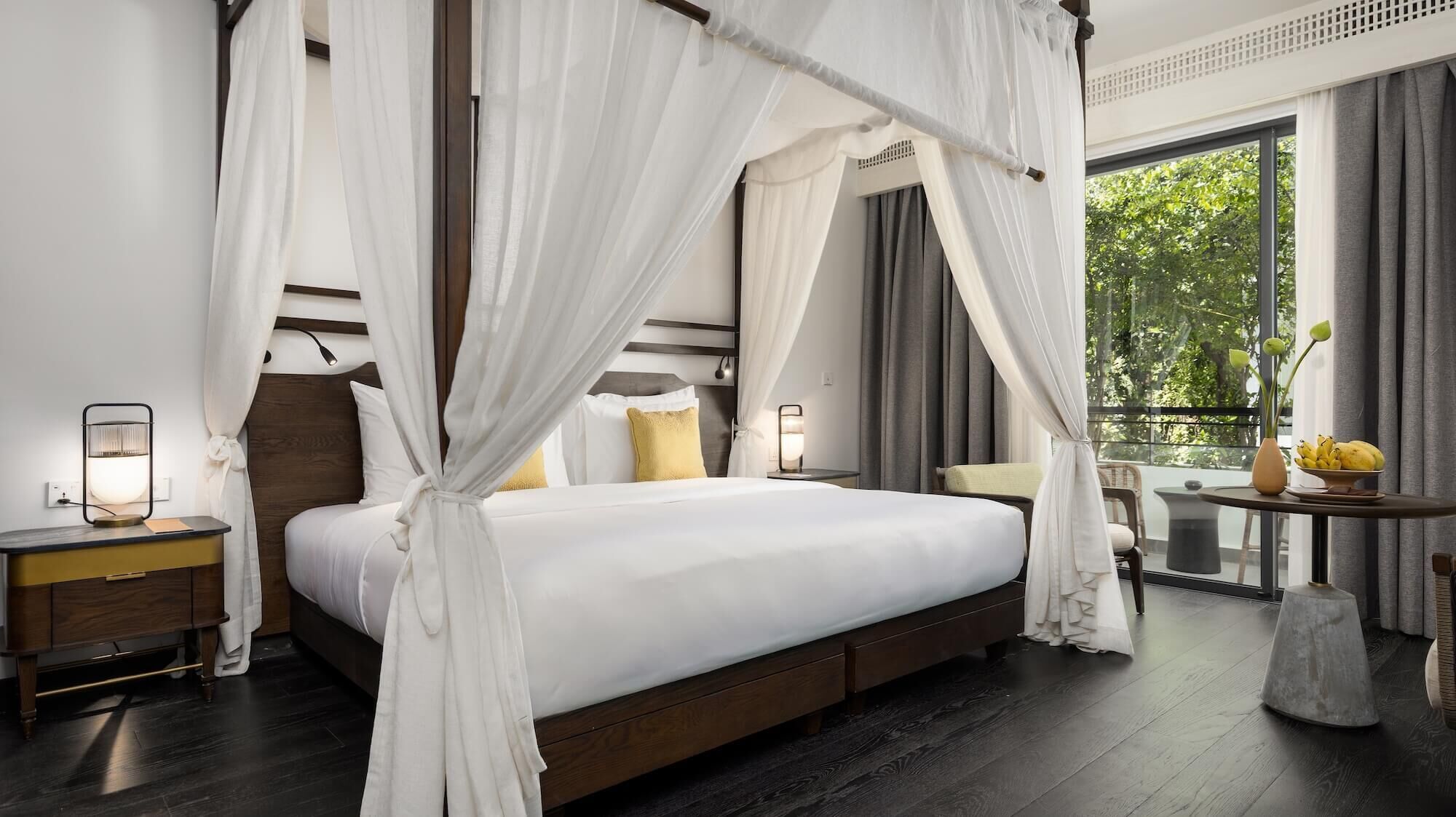 A bedroom with a four-poster bed, bedside lamps, a nightstand, and a view of greenery through the window.