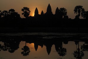 A silhouette of a temple complex against a sunset, with reflections in a calm body of water. - Anjali by Syphon (Siem Reap)