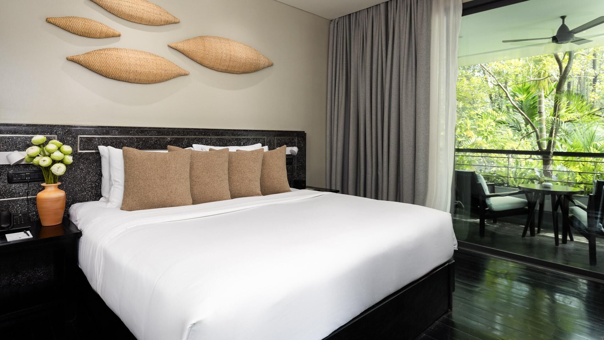 Une chambre d’hôtel avec un grand lit, une table de chevet ornée d’un vase de fleurs et une vue sur un balcon aménagé avec du mobilier d’extérieur.