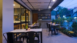 Een terras buiten met tafels en stoelen, een bar met flessen op de planken, en uitzicht op de straat.