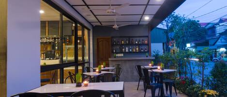 An outdoor patio area with tables and chairs, a bar with shelves of bottles, and a view of the street.