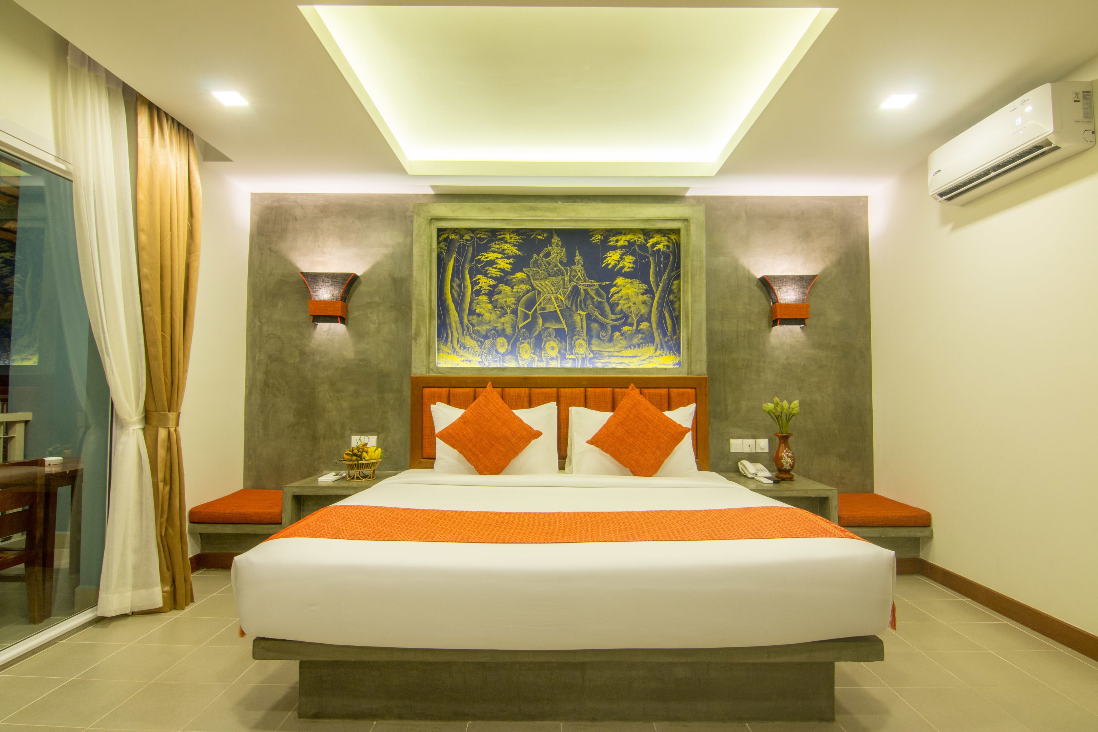 A hotel room with a large bed, two bedside tables, a nightstand, a wall-mounted air conditioner, and a large painting above the bed.