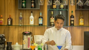 Een barruimte met een bartender die drankjes bereidt, een koffieapparaat en planken vol met verschillende flesjes drank.