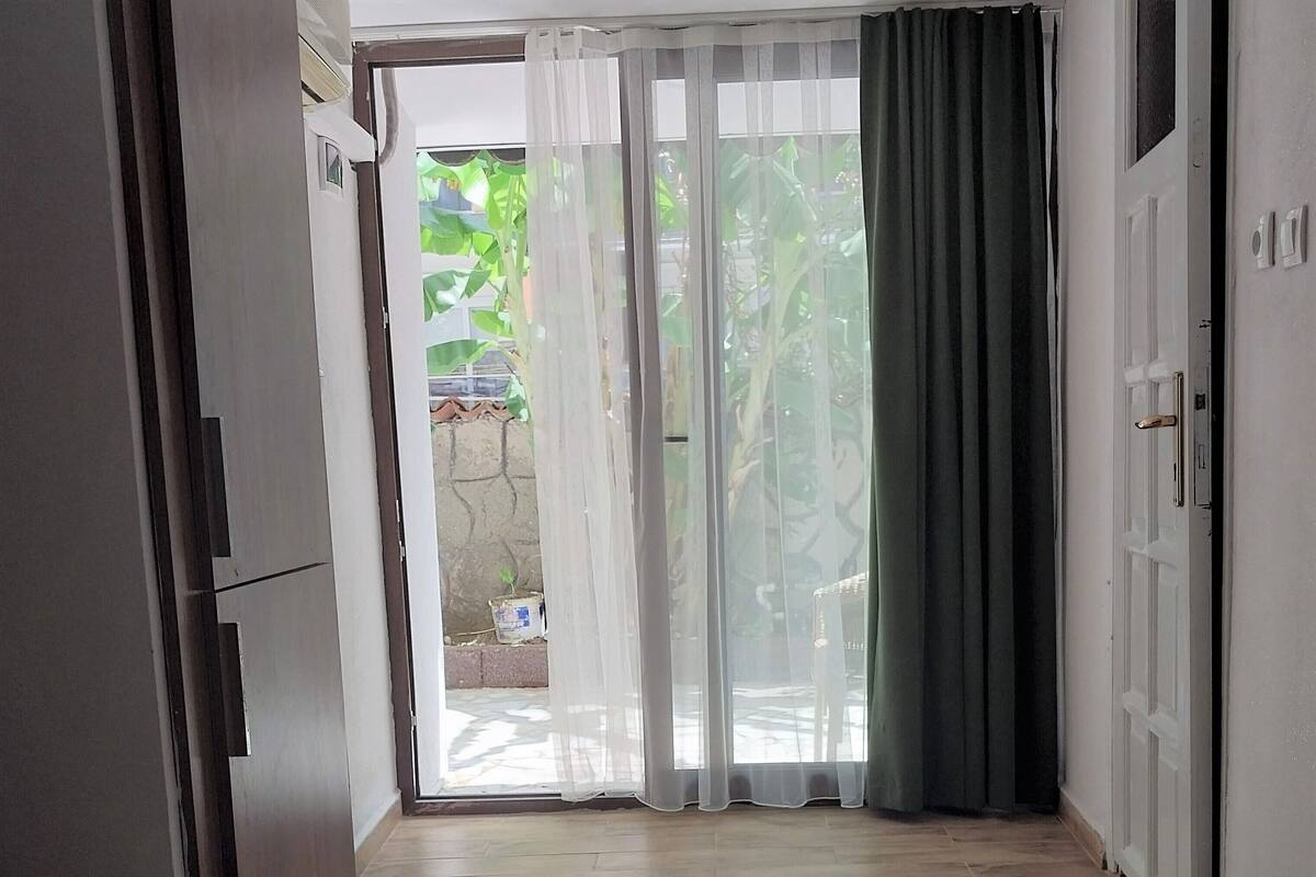 Une chambre avec un lit, un réfrigérateur, une porte et une fenêtre donnant sur l’extérieur, cette fenêtre étant dotée de rideaux.