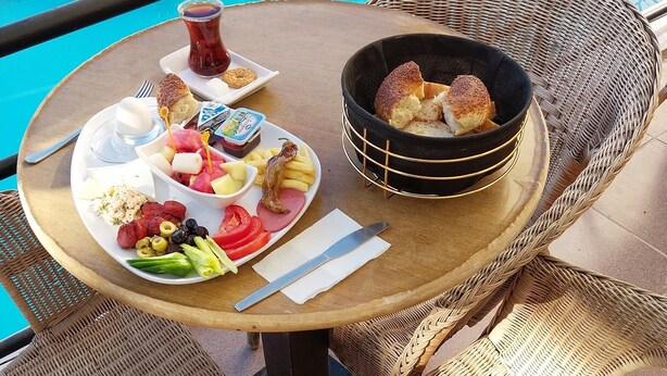 Une table à manger dressée sur un balcon, avec une variété d’aliments, notamment du pain, du fromage et des fruits, et une piscine en arrière-plan.