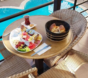 A dining table with a variety of food items including bread, cheese, and fruits, set on a balcony with a pool in the background. - Hotel Ogerim (Kusadasi)