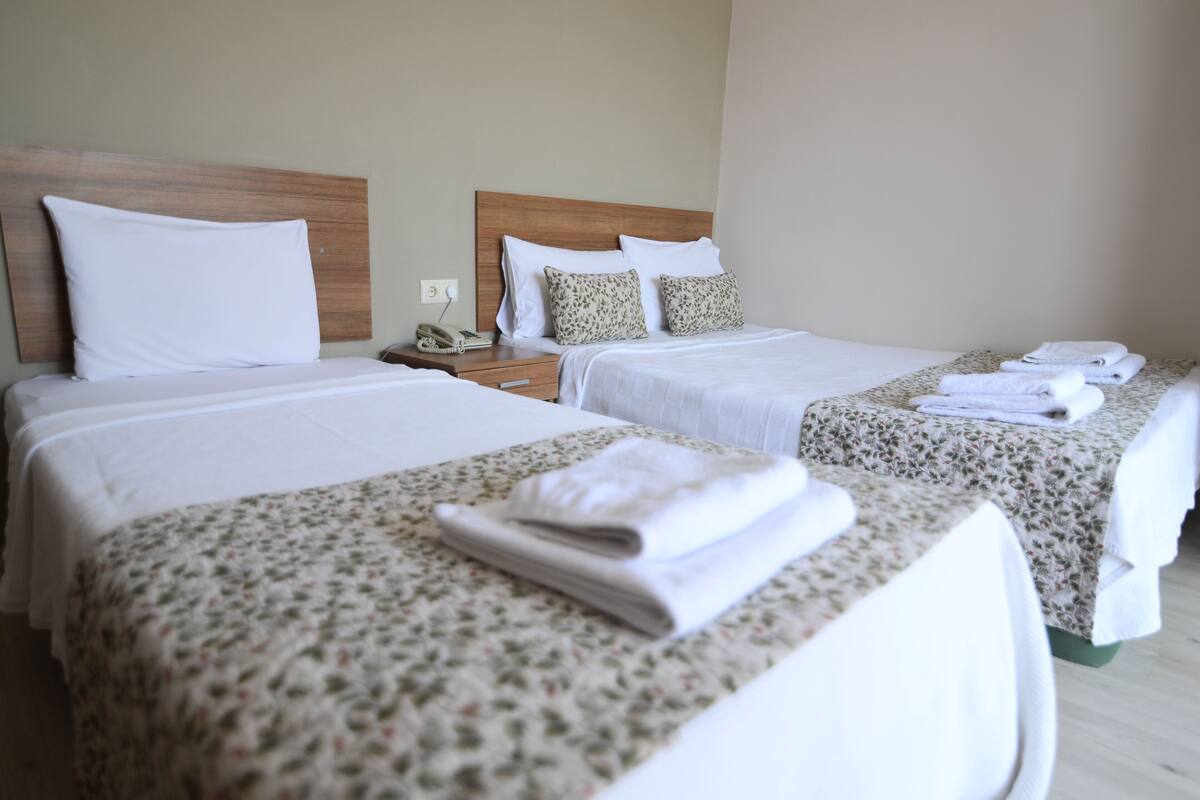 Une chambre d’hôtel avec deux lits, chacun doté d’une tête de lit en bois, de draps blancs et d’une couvre-lit à motifs floraux. Des serviettes pliées sont posées sur les lits, et un téléphone se trouve sur la table de chevet.